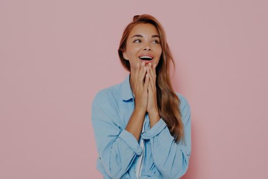 Excited woman with delightfully surprised look, touching both cheeks with hands, expresses positive emotions, wears casual blue shirt, happy to receive present or very good news. Body language