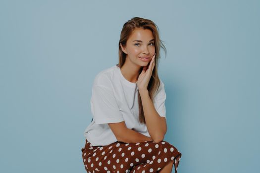 Adorable young female sincerely smiling at camera, wearing stylish outfit with polka dot skirt touching face and beilng ready for romantic date while posing on blue background. Women beauty concept