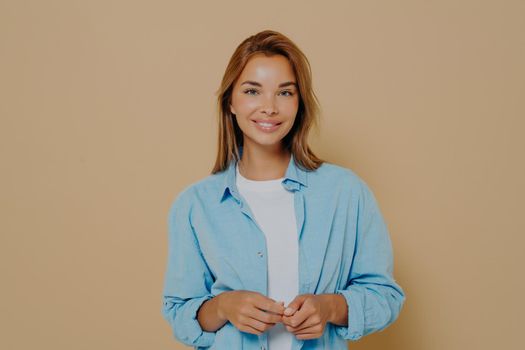 Portrait of caucasian woman in casual clothes isolated on pastel beige colored studio background, happy beautiful female model wearing blue shirt over white tshirt looks with smile at camera