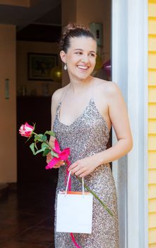 Joyful young and pretty female school graduate with stylish dress is standing on the porch smiling and holding a rose and clear presents bag.