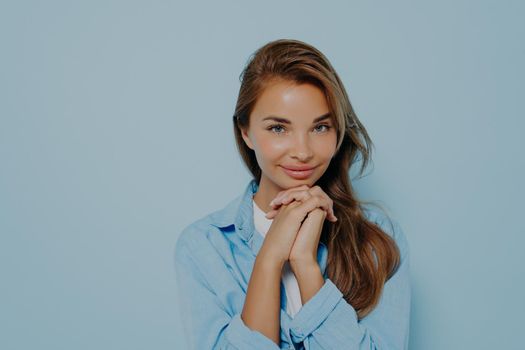 Portrait of cute caucasian young woman with intrigued face expression holding hands with palms pressed near chin and feeling excited while posing on light blue background. Copy space for text