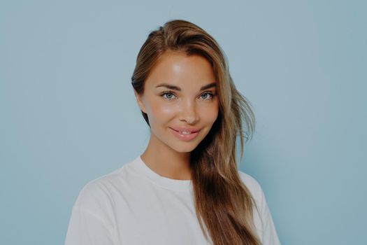 Close up portrait of smiling stylish young beautiful woman with long straight light brown hair in white t-shirt with natural light make up, isolated over blue studio background with copy space