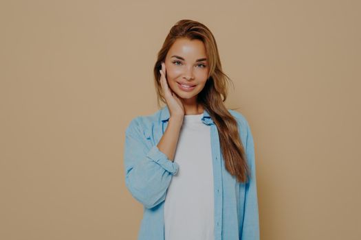 Portrait of attractive, charming brunette female with long stright hair. Wearing blue shirt over white tshirt, smiling on camera, touching her cheek with fingertips, isolated over beige background