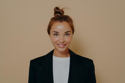 Good looking young woman with delighted face expression wearing elegant formal suit and cheerfully smiling at camera, ready for business meeting on beige background. Human face expression