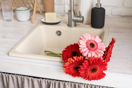 Rustic kitchen interior with white brick wall and white wooden shelves. Red and pink gerbera daisies in a kitchen sink