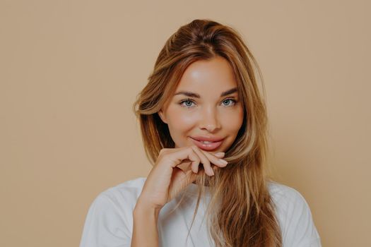 Portrait of beautiful Europian female in white tshirt looking with smile at camera holding long light brown lock of hair with fingers. Young woman with perfect healthy skin, youth and happiness