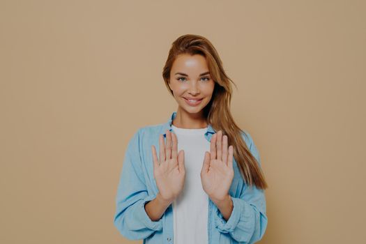 Smiling young female model with stright long hair wearing oversized blue shirt and white tshirt using hands to show stop sign while standing over beige background in studio. Positive women emotions