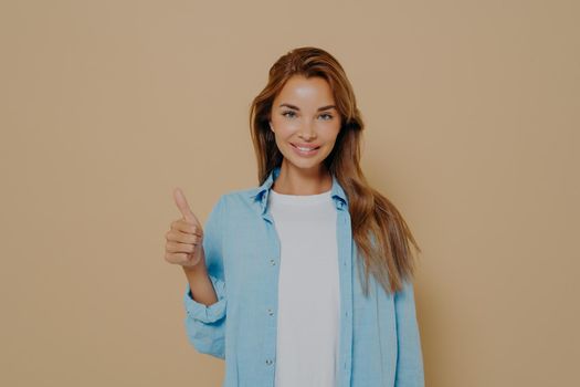 Happy young caucasian female in blue shirt with long sleeves rolled up weared over white tshirt making thumb up sign and smiling on camera with support on isolated pastel beige colored background