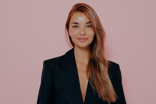 Attractive stylish business woman with long brown stright hair wears black formal jacket, looking with attentive expression and smiles at camera, her face close-up on pink background with copy space