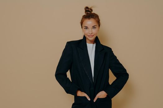 Attractive young model female in black suit with hair tied up in high bun posing with her hands in jacket pockets on a beige colored background. Fashion style studio portrait. Elegant style women