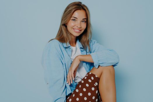 Studio portrait of beautiful young brunette woman, sitting over blue background, posing at camera and broadly smiling, wears oversized shirt and brown polka dot skirt. Beauty and fashion concept