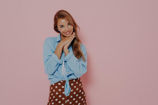 Attractive young woman in stylish outfit with beautiful sincere smile feeling love and cuteness, being shy and innocently looking at camera while standing against pink background