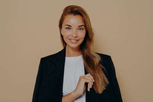 Friendly smiling brunette businesswoman with loose long hair, wears formal black jacket over white blouse, poses against beige background with blank space, happy to meet with business partners