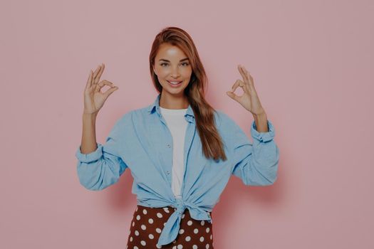 Attractive young female demonstrating agreement by gesturing okay sign with both hands or doing mudra and meditating, being calm and positive against pink background