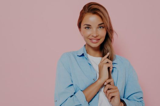 Isolated shot of attractive european student female with long straight hair looking and smiling at camera while touching her hair with hands. Positive human facial expressions and emotions