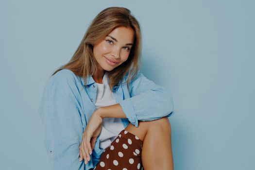Portrait of pretty smiling young brunette woman with minimal makeup in oversized shirt and polka dot skirt on blue studio background with blank space, sitting and posing with smile