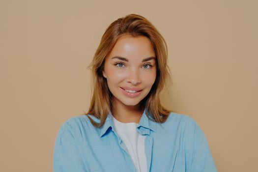 Headshot portrait of happy brunette model female in blue shirt smiling while looking at camera at studio with pastel beige background. Human facial expressions and emotions