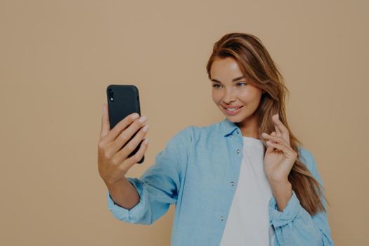 Pretty young woman makes selfie with smart phone, dressed in casual clothes, posing against beige colored studio background, uses modern technology, shares photos in social networks