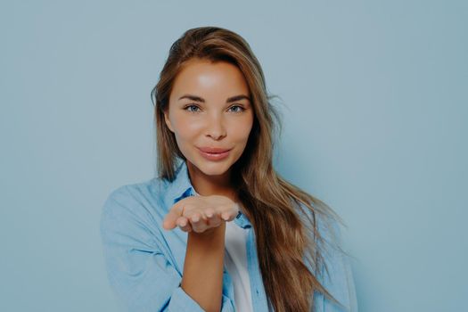 I love you. Attractive young woman with beautiful long hair in stylish outfit blowing air kiss at camera, being flirty and showing her romantic mood on light blue background. Human feeling concept