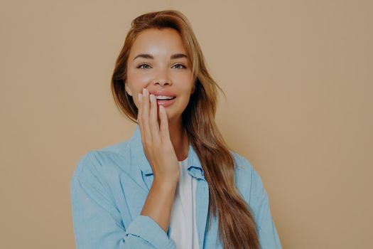 Charming tender woman touches cheek gently with hand enjoys fresh healthy skin after spa procedure has well groomed complexion looks nice in stylish blue shirt isolated on beige background. Copy space