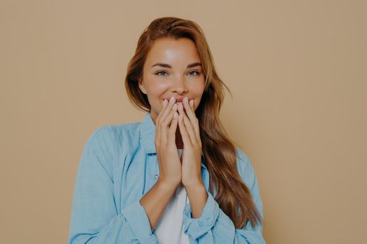 Cute young female covering mouth with hands, joyfully giggling while being shy isolated over brown background. Happy woman can not believe what she seess and looking at camera with amazed expression