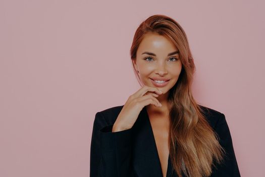 Attractive office lady with long loose stright hair and healthy skin. Smiling and lightly touches chin with fingers. Wearing stylish black blazer, looking in camera while posing over pink background