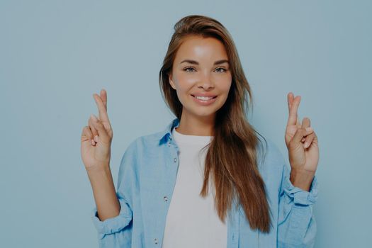 Joyful woman in casual clothes points up cross fingers asks for good luck, wishes for chance, posing on blue background studio with copy space, female student hopes for passing exam