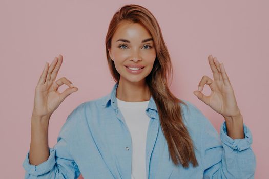 Happy young woman showing okay gesture with both hands and demonstrating aproval, looking at camera with cheerful smile, posing against pink background in stylish outfit. Body language concept