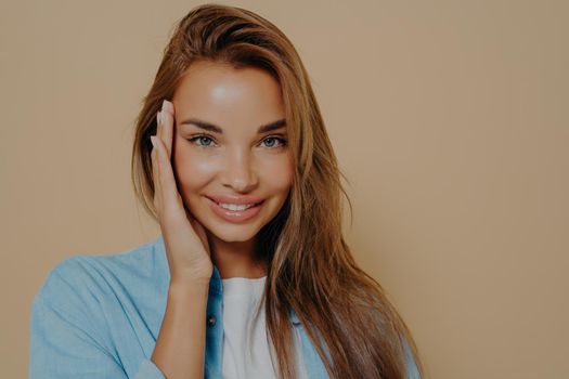 Beautiful young caucasian woman with long straight hair and healthy well cared skin, touches her face, looks positively at camera and smiling broadly, isolated on pastel beige background