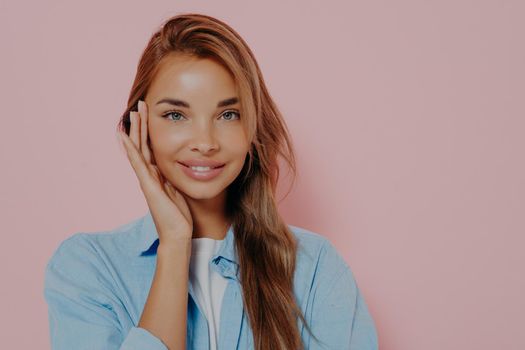 Beauty, youth and skin care concept. Headshot of attractive young woman with perfect healthy young skin, smiling at camera with happy and cheerful expression,gently touching her face with one hand