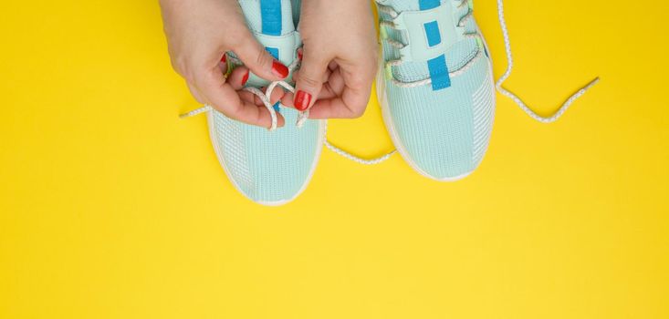 two female hands tying laces on blue textile sneakers, top view. Yellow background