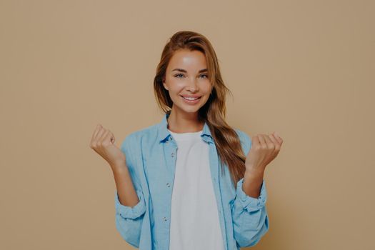 Happy young woman in stylish casual outfit holding hands in clenched fists, showing excitement and hope for best, smiling at camera while standing against beige background. People and success