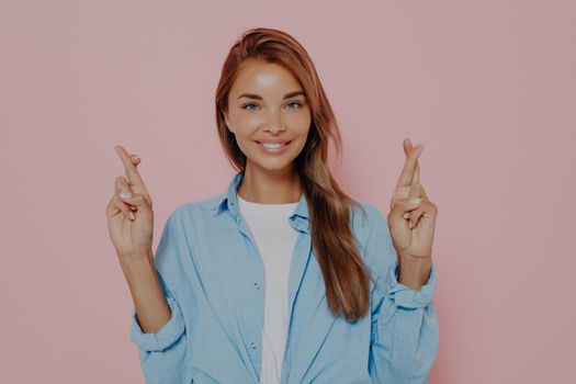 Studio shot of attractive brunette female with long stright hair having excited superstitious and joyful look, holding crossed fingers up, hoping for good luck before going for job interview or exam