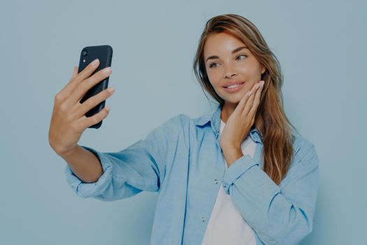Catching happy moments. Adorable cute young female with making selfie of her modern smartphone, gently smiling at camera while posing isolated on light blue background
