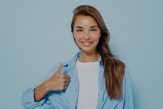 Happy young caucasian female wearing blue long sleeved shirt making thumb up sign and smiling cheerfully, showing her support and respect to someone. Body language. I like that. Good job
