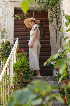 Beautiful female tourist wearing big straw sun hat and sundress standing on old vintage stone house staircase during summer travel on Mediterranean cost on hot summer day.