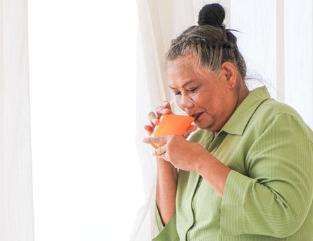 Senior Asian women Drinking orange juice standing by the window in the background of the room, the concept of living the elderly, retirement and healthy lifestyle.