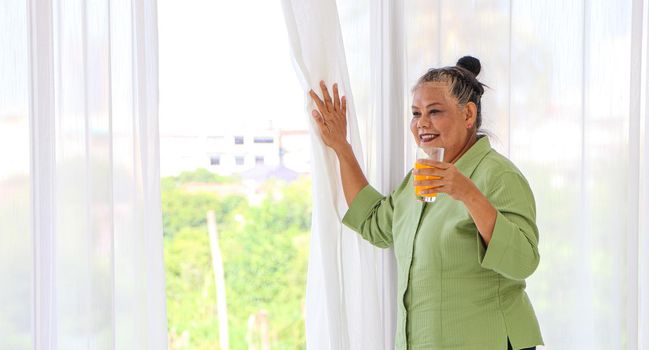 Senior Asian women Drinking orange juice standing by the window in the background of the room, the concept of living the elderly, retirement and healthy lifestyle.