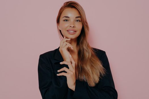 Tender young woman with long straight hair has healthy well cared skin, gently touches face, looks positively at camera, wears black classic blazer, isolated on pink background. Women beauty concept