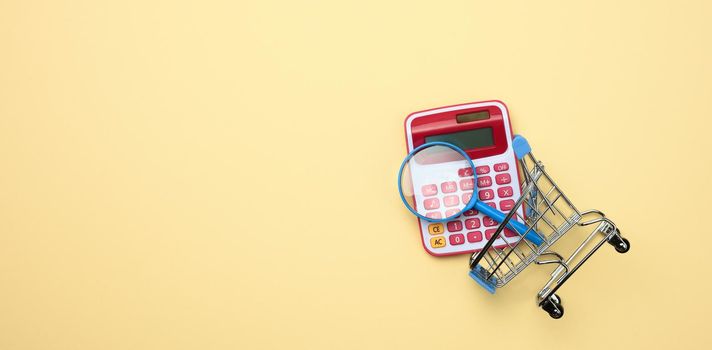 pink plastic calculator in a miniature pink trolley on a yellow background. Budget planning concept, savings counting, cost control, flat lay