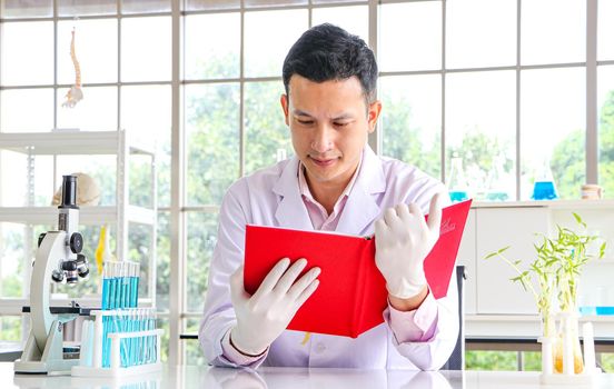 A scientist or researcher in an Asian male wearing a white coat is working and reading a book to find information. Being in a scientific laboratory With test tube equipment