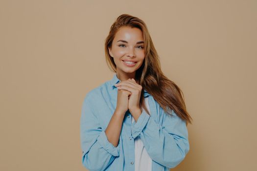 Beauty and positive emotion concept. Charming young smiling european woman keeps hands near face smiles tenderly at camera dressed in casual wear, poses against beige studio background