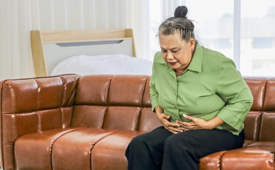 Asian elderly woman Holding the stomach in pain while sitting on the sofa in the room Health care concept Women suffering from abdominal pain Senior female health problems and people's concepts