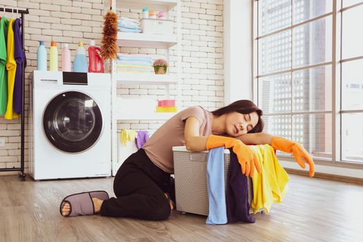 A cute Asian woman wearing a brown shirt. She is sleeping beside a laundry basket in the laundry room due to chores from doing housework and washing clothes during the day.