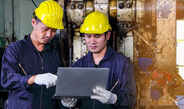 Two skilled Asian workers, dressed in helmet uniforms, are using a computer to analyze the metal press maintenance inspection data. The concept of teamwork at the factory