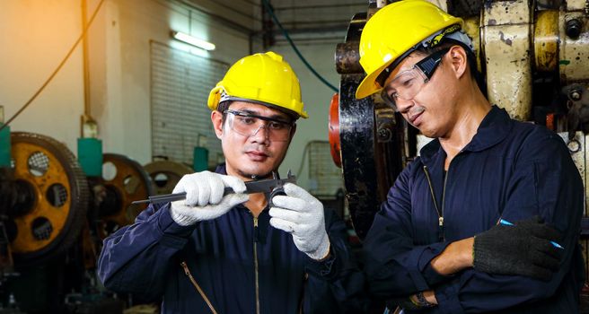 Two Asian male workers in safety clothing use Vernier calipers. Inspect the quality and measure the spare parts of working machines in industrial plants and production processes.