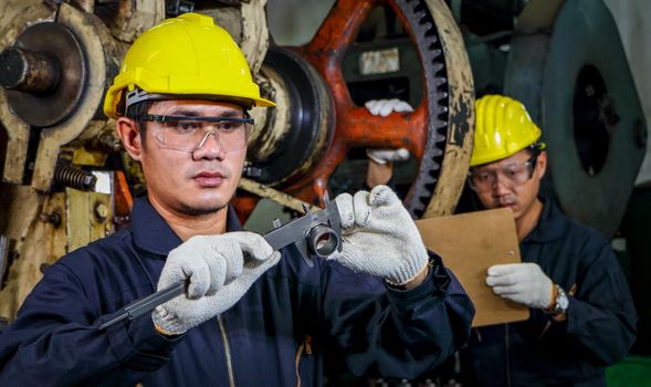 Two Asian male workers in safety clothing use Vernier calipers. Inspect the quality and measure the spare parts of working machines in industrial plants and production processes.