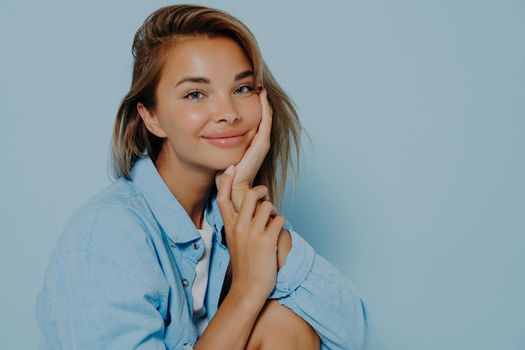 Positive caucasian woman with clear skin and beautiful long hair, leaning head on hand while smiling happily at camera, expressing positivity and happiness while posing on light blue background