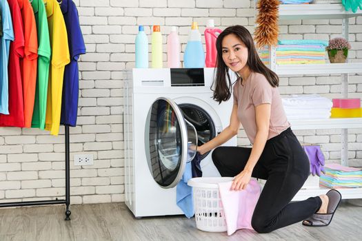 A smiling and cute Asian woman carrying a laundry basket and posing beside the washing machine. Picking up clothes in the washing machine. There are empty shelves and clothes racks.