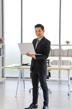 An Asian business man stands and holds a laptop computer to work at the office. Use apps for social media and online technology news for business strategies. Smile and be happy